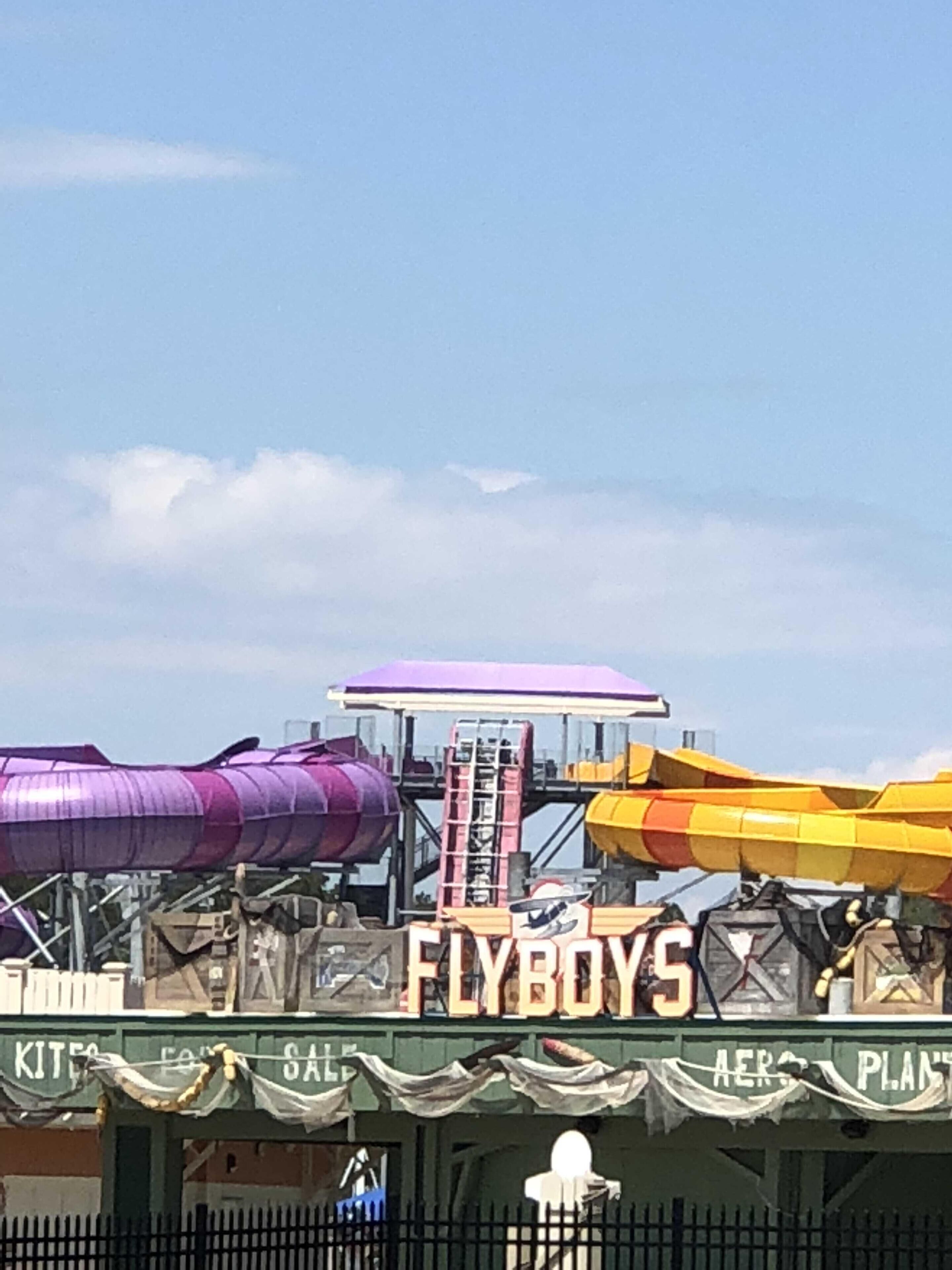 Una atracción acuática con un tobogán púrpura y amarillo, una plataforma con el rótulo 'FLYBOYS' y una valla en primer plano.