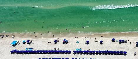 Une vue aérienne d’une plage, avec des rangées de parasols bleus et de chaises longues, ce qui indique qu’il s’agit d’un complexe hôtelier balnéaire.