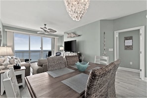 A spacious living room with a large dining table, comfortable seating, a ceiling fan, and a view of the ocean through large windows.