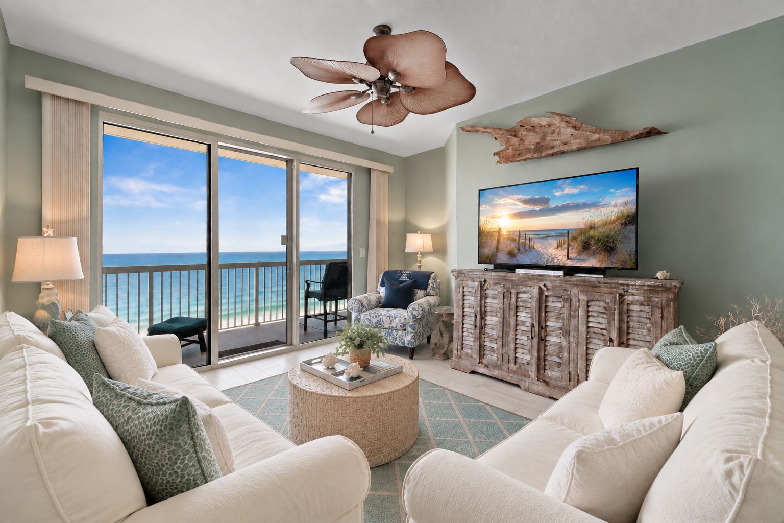 A living room with a view of the ocean, a ceiling fan, a television, and a wooden cabinet.