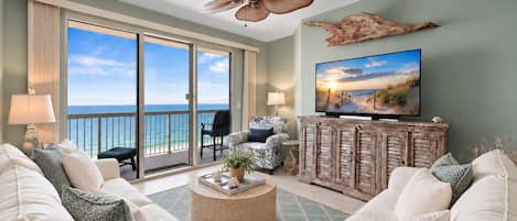 A spacious living room with a large TV, comfortable seating, and a view of the ocean through the sliding doors.
