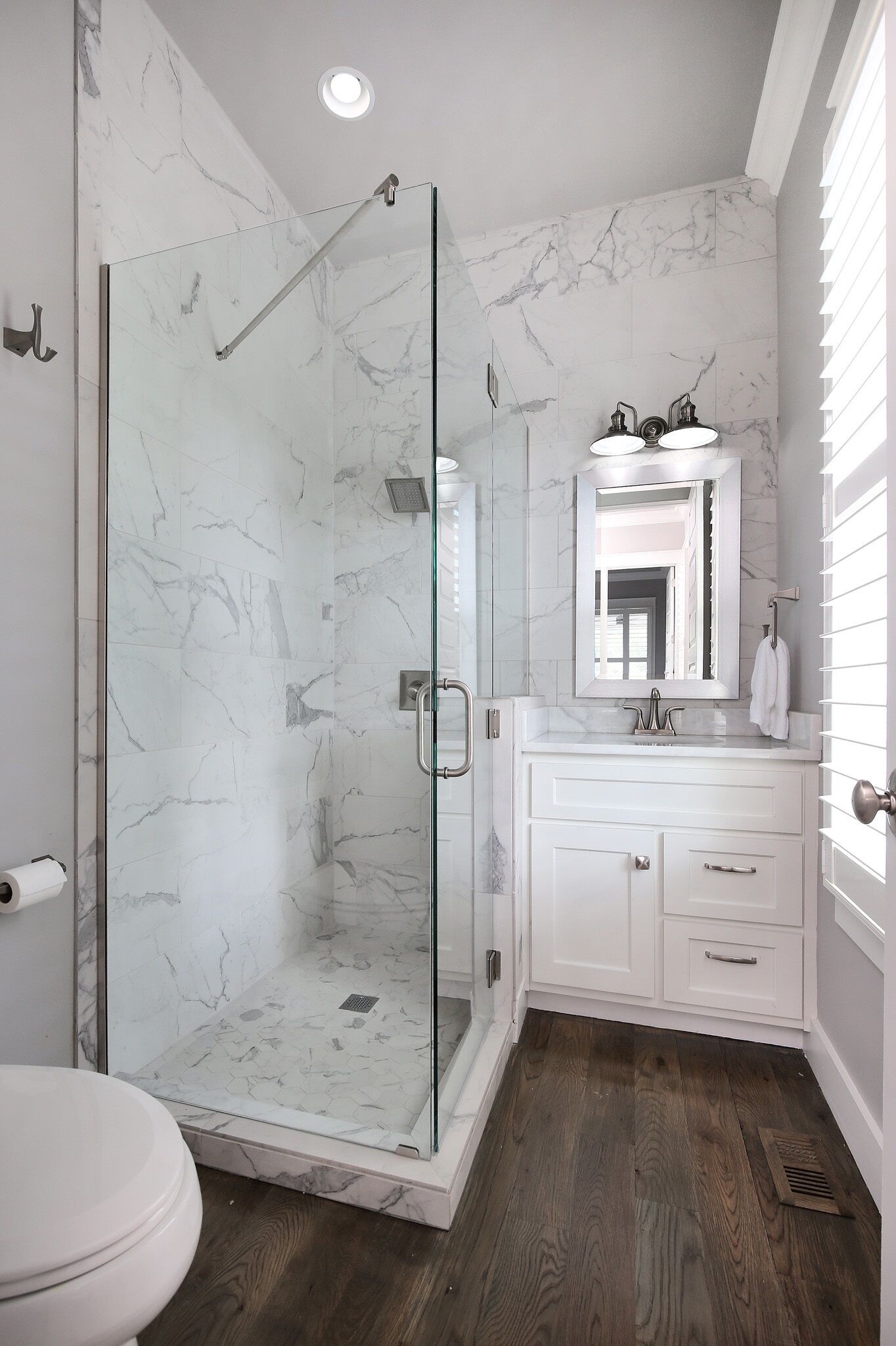 A bathroom with a glass-enclosed shower, white marble walls, a white vanity with a mirror, and a wooden floor.