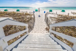 Una escalera de madera que baja a una playa arenosa, con una valla de madera y sillas verdes.