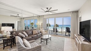 A spacious living room with a large sofa, a coffee table, and a TV. The room has large windows offering a view of the beach and ocean. - Oceanfront Elegance, Great Views, on the beach, steps to the sand 2BR/2BA (Destin)