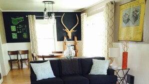 Ein Wohnzimmer mit einer schwarzen Couch, zwei kleinen Tischen, einem Kuhfellteppich, Holzboden, einer Chandelieber und einer Dekoration aus einem Hirschschädel.