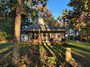 A small wooden cabin with a porch, surrounded by trees and a grassy yard.