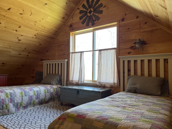A cozy attic bedroom with two beds, a window with curtains, a wooden ceiling, and a decorative clock.