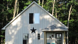 A small, single-story wooden house with a gabled roof, a large star decoration, and a porch with two chairs. - Grey Goose Cottage In The Heart Of The FingerLakes (Keuka Lake Penn Yan)