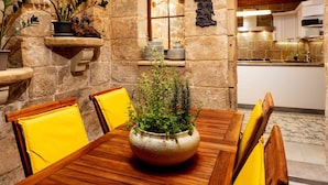 A stone-walled dining area with a wooden table and yellow chairs, a potted plant, and kitchen cabinets in the background. - The 3Cities Auberge (Cospicua)