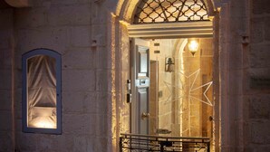 A stone building entrance with an arched doorway, a metal gate, and a small window with a curtain. - The 3Cities Auberge (Cospicua)