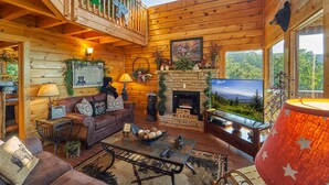 A cozy cabin interior with a fireplace, wooden walls, a balcony with plants, and a large window offering a scenic view.