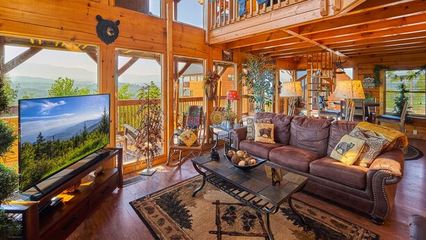 A spacious wooden cabin with a large TV, leather sofa, and a balcony with a bear decoration.