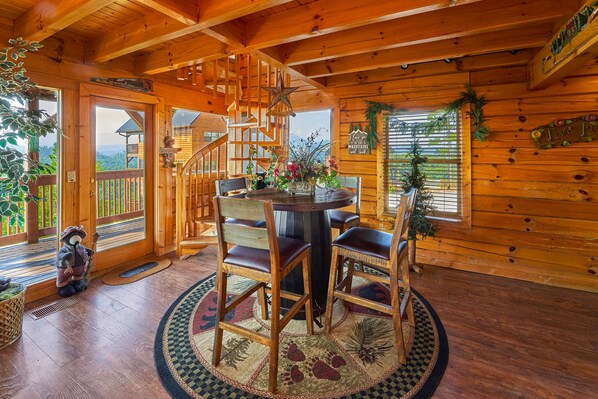 Interior de una cabaña de madera con comedor, que incluye una mesa redonda y sillas, una escalera y vistas a través de las ventanas.
