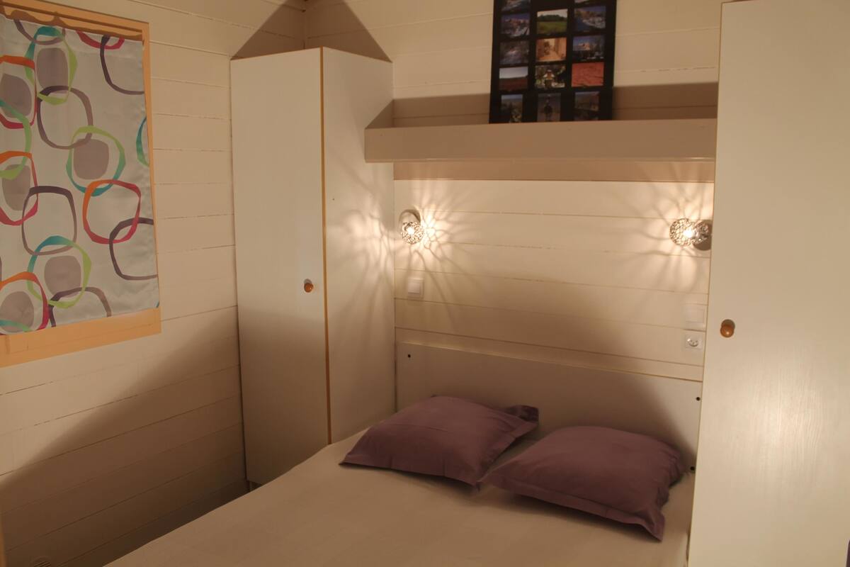 A small, compact room with a bunk bed, a wardrobe, and a wall-mounted shelf with pictures.