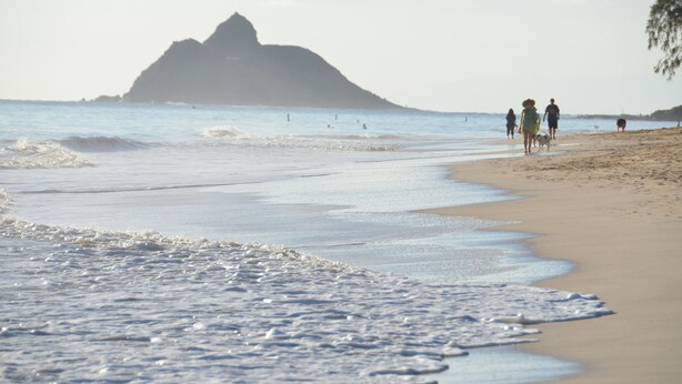 A beach with waves, a distant mountain, and people walking.