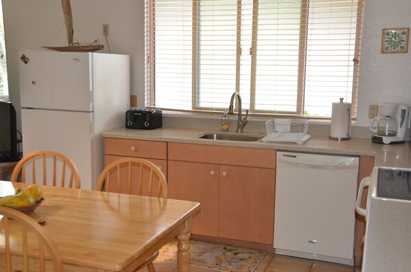 A compact kitchen with a sink, a dishwasher, a refrigerator, and a microwave. There is a dining table with chairs and a window with blinds. - Steps to the Beach Guesthouse (Kailua)