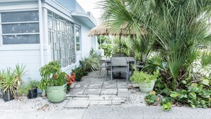 A house with a patio area featuring a table and chairs, surrounded by various potted plants and a palm tree.