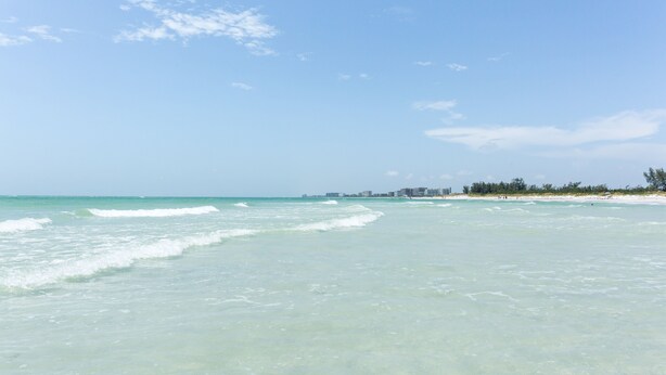 A beach with clear turquoise water, a sandy shore, and a distant city skyline.