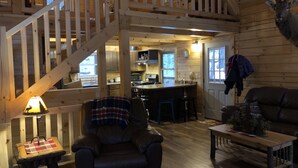 Interior acogedor de una cabaña con una escalera de madera, comedor, cocina y sala de estar con un sillón de cuero y una mesita de centro.