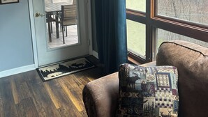A room with a wooden floor, a brown leather couch with a patterned pillow, a door leading to a patio with a table and chairs, and a view of mountains through a window with blue curtains. - Right beside pool (Gatlinburg)