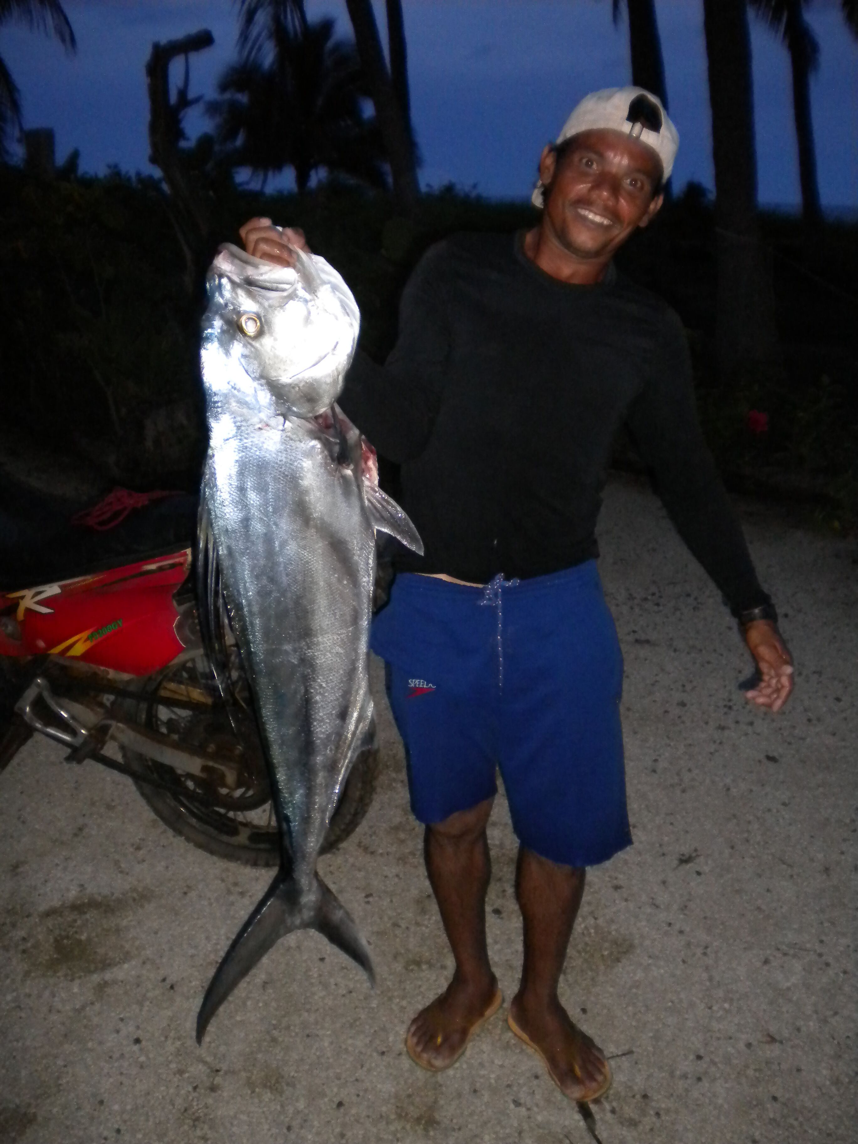 Una persona tiene in mano un pesce grande, in piedi accanto a una motocicletta su una superficie asfaltata.