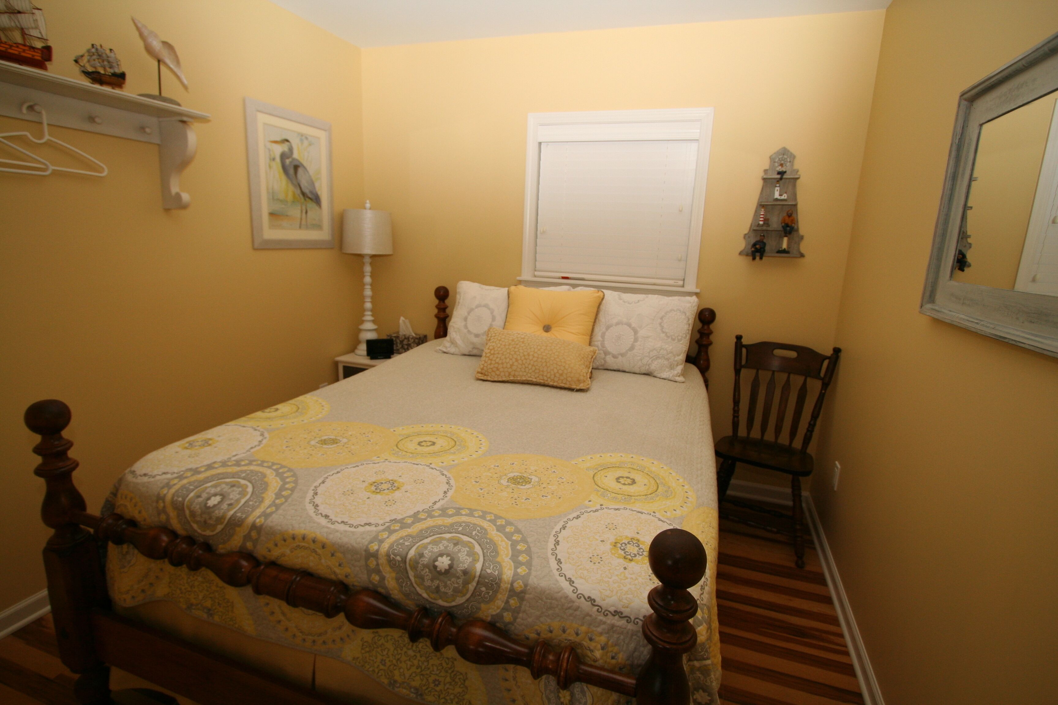 A bedroom with a bed, a chair, a nightstand, a lamp, a wall-mounted shelf with decorative items, and a window with a blind.