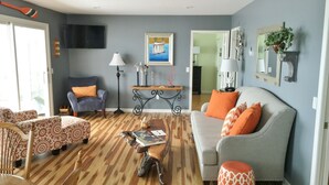 Ein Wohnzimmer mit grauem Sofa, gemusterten Sesseln, einem Holztisch und einem an der Wand montierten Fernseher.