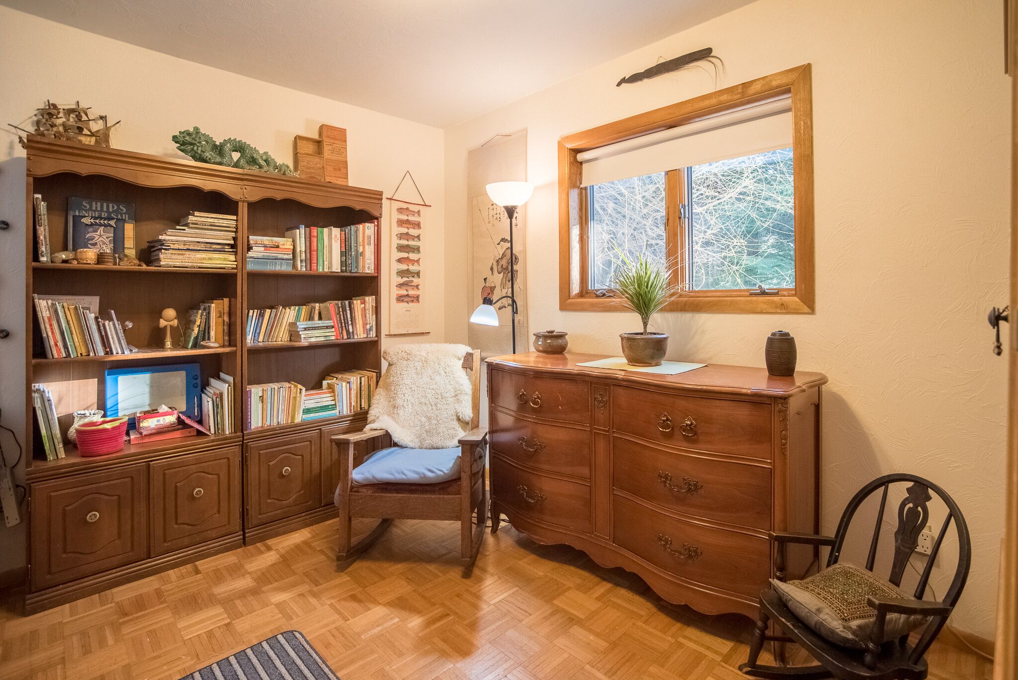 Un rincón acogedor para leer con una estantería de madera, una silla, un escritorio con lámpara y una ventana con vistas a árboles.