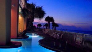 A swimming pool with a curved edge, surrounded by palm trees and a building with a balcony.