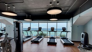 A gym with treadmills, exercise bikes, and a rowing machine, overlooking a beach.