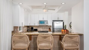A modern kitchen with white cabinets, a stainless steel refrigerator, a microwave, and a ceiling fan. There are three wicker chairs at the bar counter. - Gorgeous second floor, ocean front with huge wrap around balcony!!! (Providenciales)
