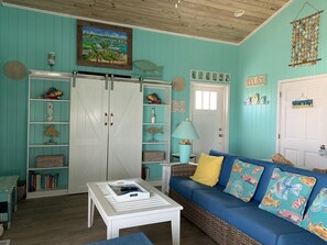 A room with teal walls, a white door, a blue sofa with colorful cushions, a white coffee table, and a bookshelf with decorative items. - Ocean Views, Sea Breezes and Pool -Family Friendly 3BR/2BA- CATCH A WAVE BAHAMAS (Hope Town)