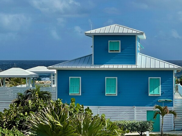 A blue building with a white roof and green shutters, surrounded by lush greenery and a view of the ocean. - Ocean Views, Sea Breezes and Pool -Family Friendly 3BR/2BA- CATCH A WAVE BAHAMAS (Hope Town)