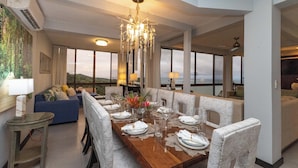 A dining area with a long table set for six, surrounded by upholstered chairs. A chandelier hangs above, and the room has large windows offering a view of the outdoors. - Villa Tortuga: Ocean Views Chef Included, Daily Housekeeping & Concierge Service (Quepos)