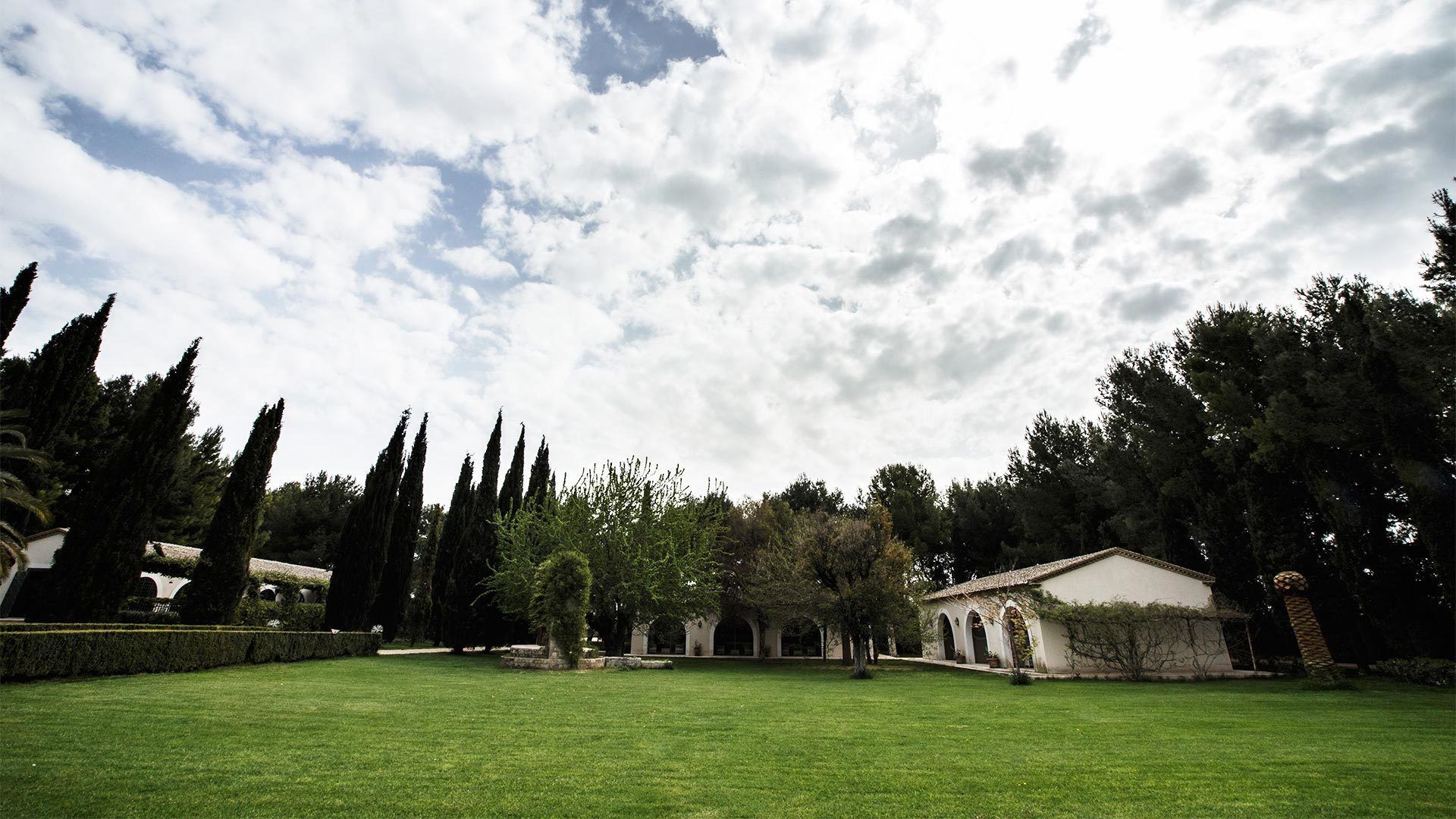 A spacious courtyard with a well-maintained lawn, tall cypress trees, and a building with arched windows and a tiled roof.