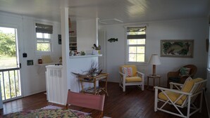 Una habitación acogedora con pisos de madera, una mesa de comedor con mantel floral, dos sillas de mimbre, una mesita auxiliar con lámpara y una ventana con contraventanas.