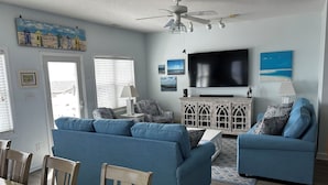 A living room with a blue sofa set, wooden dining table, and chairs, a TV, and a ceiling fan.