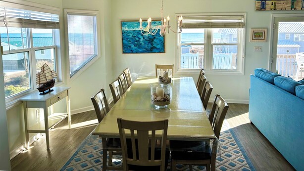 A dining area with a long table, chairs, a chandelier, and a view of the ocean through large windows.