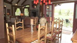 Une table à manger en bois, des chaises, un lustre et une cheminée dans une pièce dotée de poutres apparentes et de grandes fenêtres.