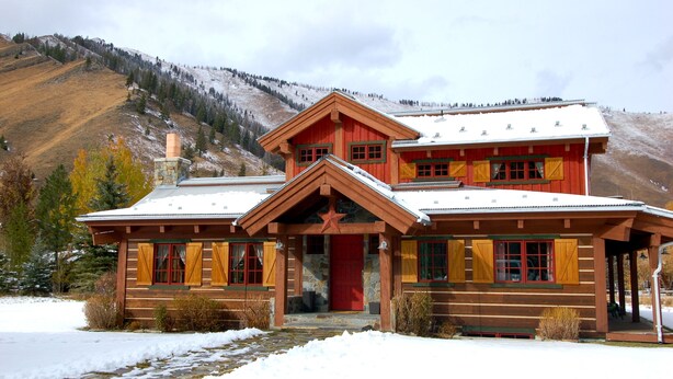 Une cabane en rondins avec une porte rouge et de nombreuses fenêtres, entourée d’un paysage enneigé et dominée par un décor montagneux.