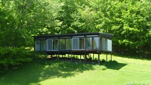 A modern houseboat with a glass facade, elevated on stilts over a body of water, surrounded by lush greenery. - Family-Friendly Home With Incredible Berkshire Views And A Private Swim Pond (Stephentown)