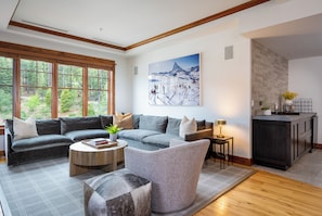 Un salon moderne avec un canapé d’angle, une table basse ronde et un fauteuil gris. Un tableau représentant un paysage enneigé est accroché au mur.