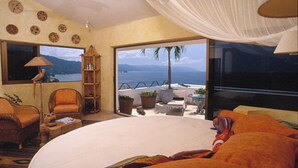 Ein Schlafzimmer mit einem großen Bett, einem Sessel, einem kleinen Tisch, einer Lampe und Meerblick durch große Fenster.