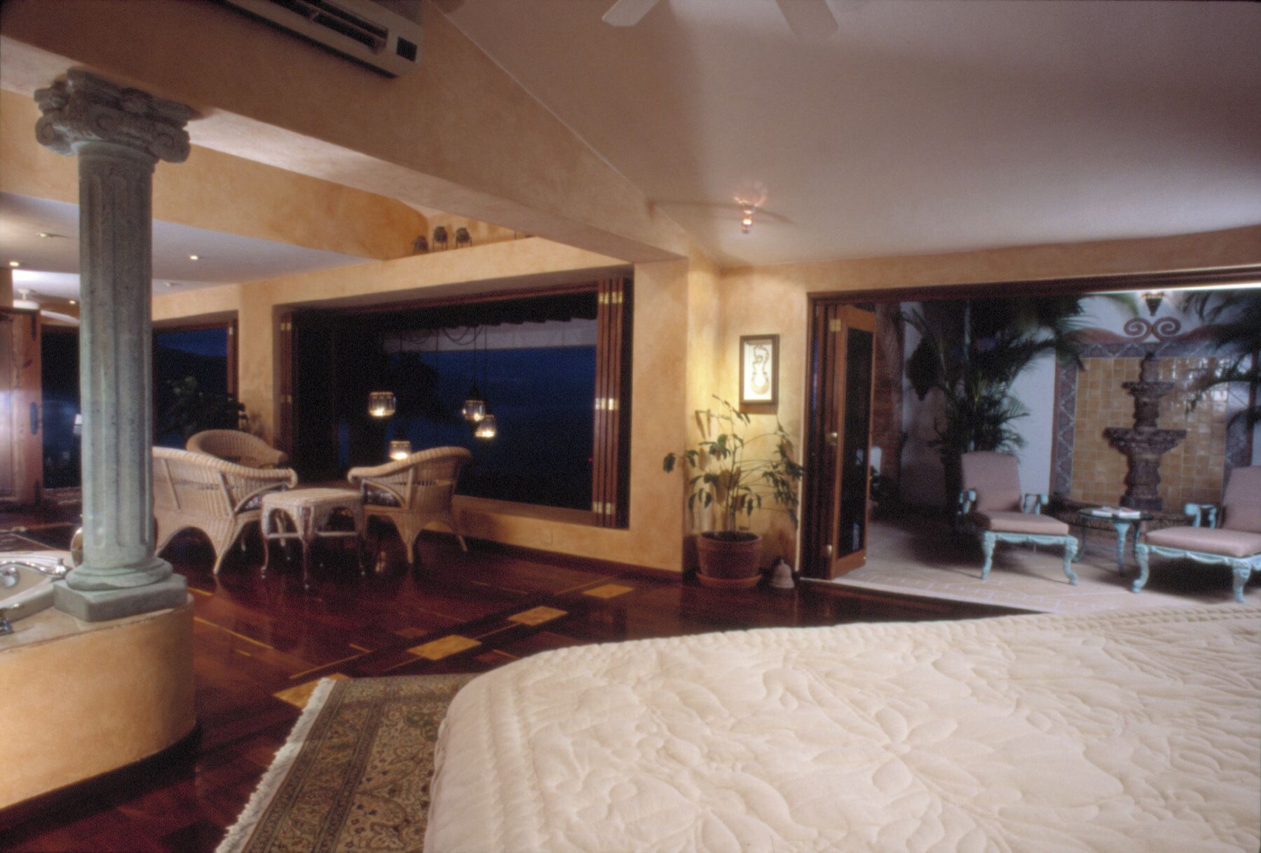 A spacious bedroom with a large bed, a sitting area with chairs, a potted plant, and a view through an open door.