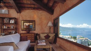 Ein gemütliches Wohnzimmer mit einem weißen Sofa, einem Korbtisch und einem Bücherregal. An der Wand befinden sich ein großer Spiegel und eine Lampe. Das Zimmer bietet einen Blick auf das Meer und Gebäude.