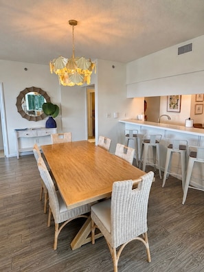 A dining area with a wooden table and wicker chairs, a pendant light, and a kitchen with white bar stools.