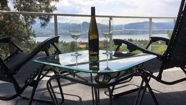A glass table with a bottle and two wine glasses, overlooking a lake and mountains.