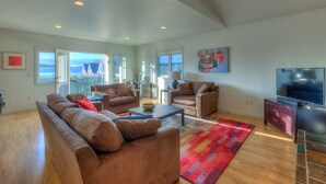 A spacious living room with a large sofa set, a coffee table, and a TV. There are multiple windows providing natural light and a view of the outdoors. - Water view! 
Luxury Port Suite -- walk to cafes, shops, galleries & beaches. (Eastsound)