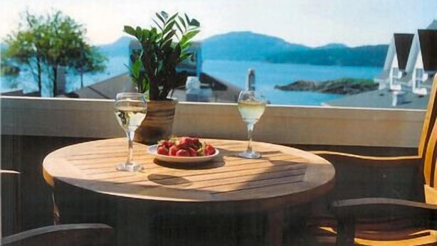 A round wooden table with a plate of food and two glasses of white wine, set against a scenic view of a body of water and mountains. - Water view! 
Luxury Port Suite -- walk to cafes, shops, galleries & beaches. (Eastsound)