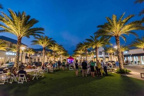 A nighttime outdoor area with palm trees, tables, and chairs, and a stage with people. - Gulf views Beach House & Large Private Pool, 2 owner suites; Walk 2 Beach (Panama City Beach)